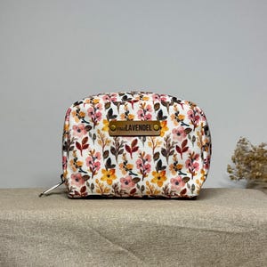 Könnte beinhalten: Eine weiße Reißverschlusstasche mit einem Blumendruck in Rosa-, Gelb- und Brauntönen. Die Tasche hat ein braunes Leder-Etikett mit dem Text "Paula LAVENDEL" darauf.