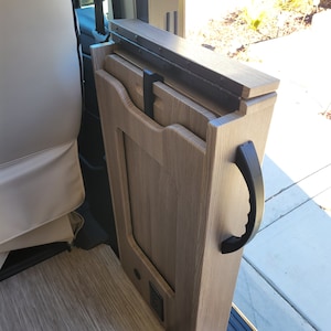 May include: A wooden folding table with a black handle, stored in a camper van. The table is made of light wood with a wood grain pattern. The table is folded up and stored in a vertical position.