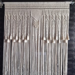 May include: Cream-colored macrame curtain with intricate woven patterns and hanging tassels. The curtain is suspended from a metal rod, showcasing a bohemian style. The design features geometric shapes and knotted details, adding texture and visual interest to the textile.