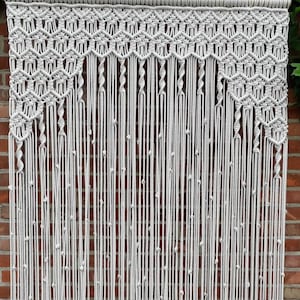 Pode incluir: Uma cortina de macramé branca com um padrão geométrico. A cortina é feita de cordão de algodão grosso e pendura em uma haste de metal.