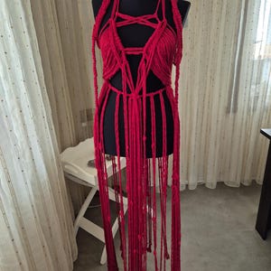 May include: A vibrant red macrame garment featuring an intricate knotted bodice with a pentagram-inspired design, thick braided shoulder straps, and long, dangling fringe cords that extend downward from the waist, displayed on a black mannequin.