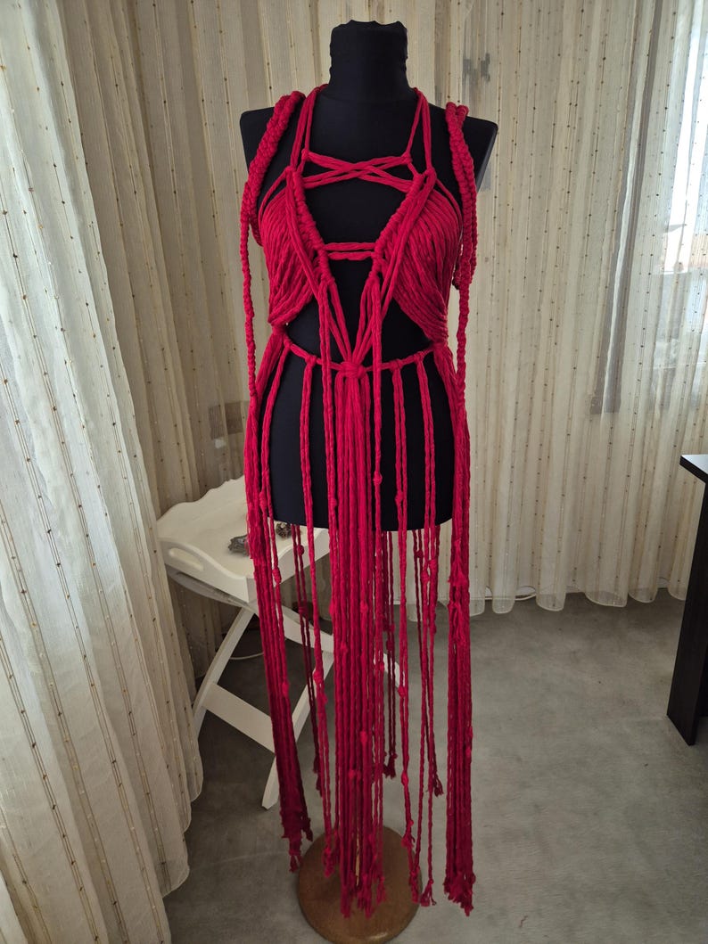May include: A vibrant red macrame garment featuring an intricate knotted bodice with a pentagram-inspired design, thick braided shoulder straps, and long, dangling fringe cords that extend downward from the waist, displayed on a black mannequin.
