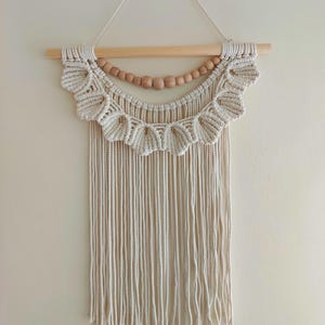 May include: A macrame wall hanging featuring a natural, off-white color scheme. The design includes a scalloped top section, a row of wooden beads, and long, fringed strands. It is suspended from a light-colored wooden dowel.