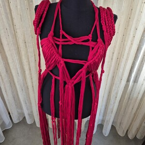 May include: A vibrant red macrame top crafted from thick cotton cord, featuring an intricate knotted bodice with a deep neckline, braided shoulder straps, and long, dangling fringe elements that extend downward from the waist area.