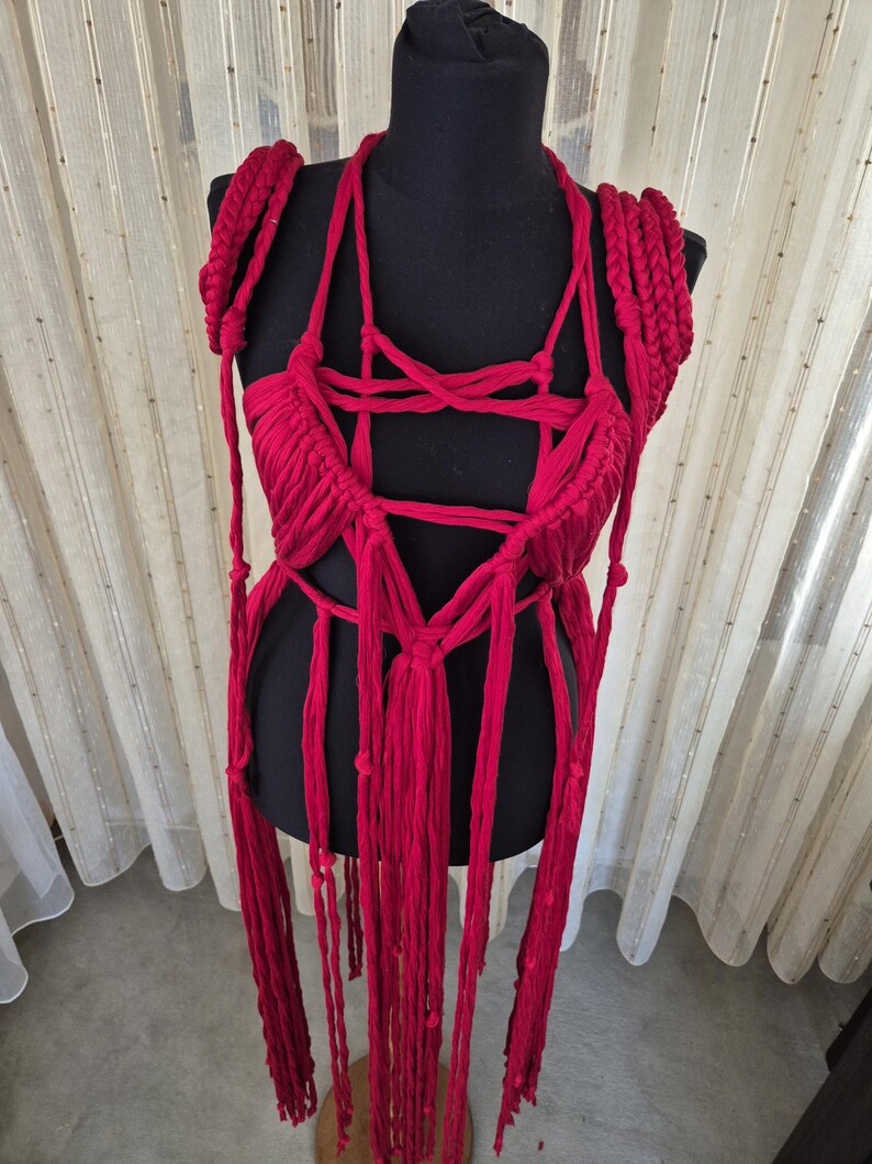 May include: A vibrant red macrame top crafted from thick cotton cord, featuring an intricate knotted bodice with a deep neckline, braided shoulder straps, and long, dangling fringe elements that extend downward from the waist area.