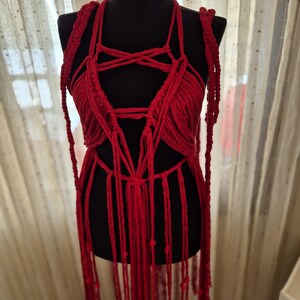 May include: A vibrant red macrame top featuring intricate knotted cord patterns, a halter-style neckline, and long, dangling fringe elements that extend downward. The garment is constructed from thick, textured cotton-like cord in a deep red hue.