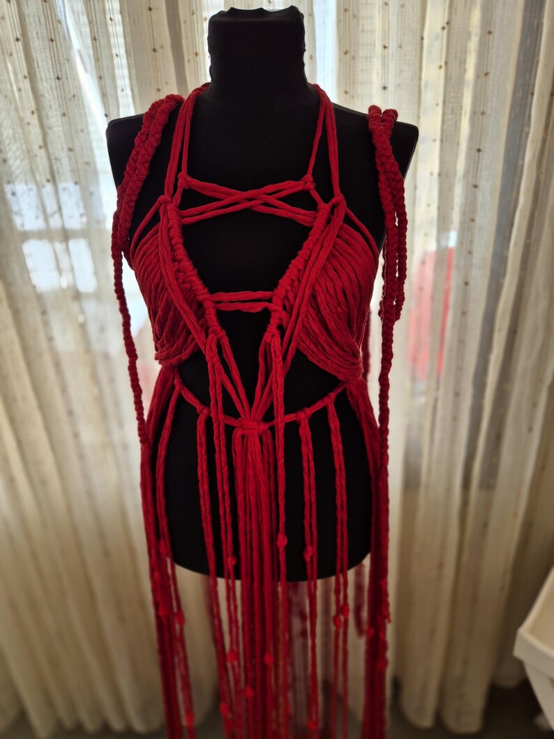 May include: A vibrant red macrame top featuring intricate knotted cord patterns, a halter-style neckline, and long, dangling fringe elements that extend downward. The garment is constructed from thick, textured cotton-like cord in a deep red hue.