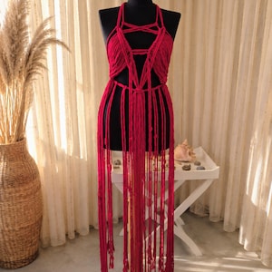 May include: A vibrant red macrame halter top featuring an intricate knotted design with a deep neckline and long, flowing fringe that extends past the waist, displayed on a black mannequin torso to showcase the detailed cord work and texture.