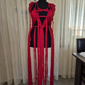 May include: A vibrant red macrame garment featuring an intricate knotted bodice with a deep V-neckline and long, flowing fringe strands that extend downward. The piece is constructed from thick, soft cord and is displayed on a black mannequin torso.