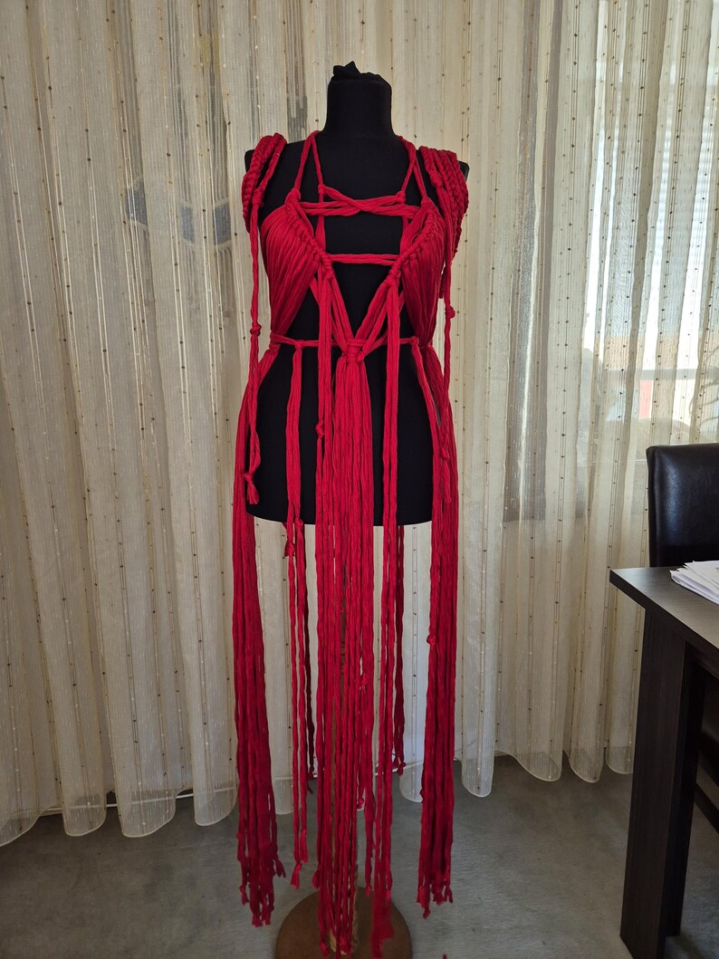 May include: A vibrant red macrame garment featuring an intricate knotted bodice with a deep V-neckline and long, flowing fringe strands that extend downward. The piece is constructed from thick, soft cord and is displayed on a black mannequin torso.