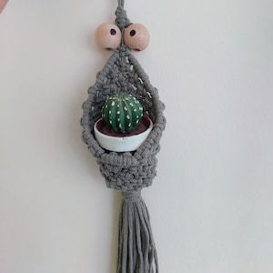 Könnte beinhalten: Ein grauer Makramee-Pflanzenaufhänger mit einem kleinen grünen Kaktus in einem weißen Topf. Der Aufhänger hat Holzperlen und eine Quaste. Der Kaktus hat weiße Punkte. Der Pflanzenaufhänger wird von einer grauen Schnur gehalten.