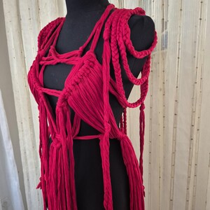 May include: A vibrant red macrame garment featuring intricate hand-knotted cord patterns, thick braided shoulder straps, and long dangling fringe elements. The piece is constructed from soft fabric strips, creating a complex, open-weave structure.