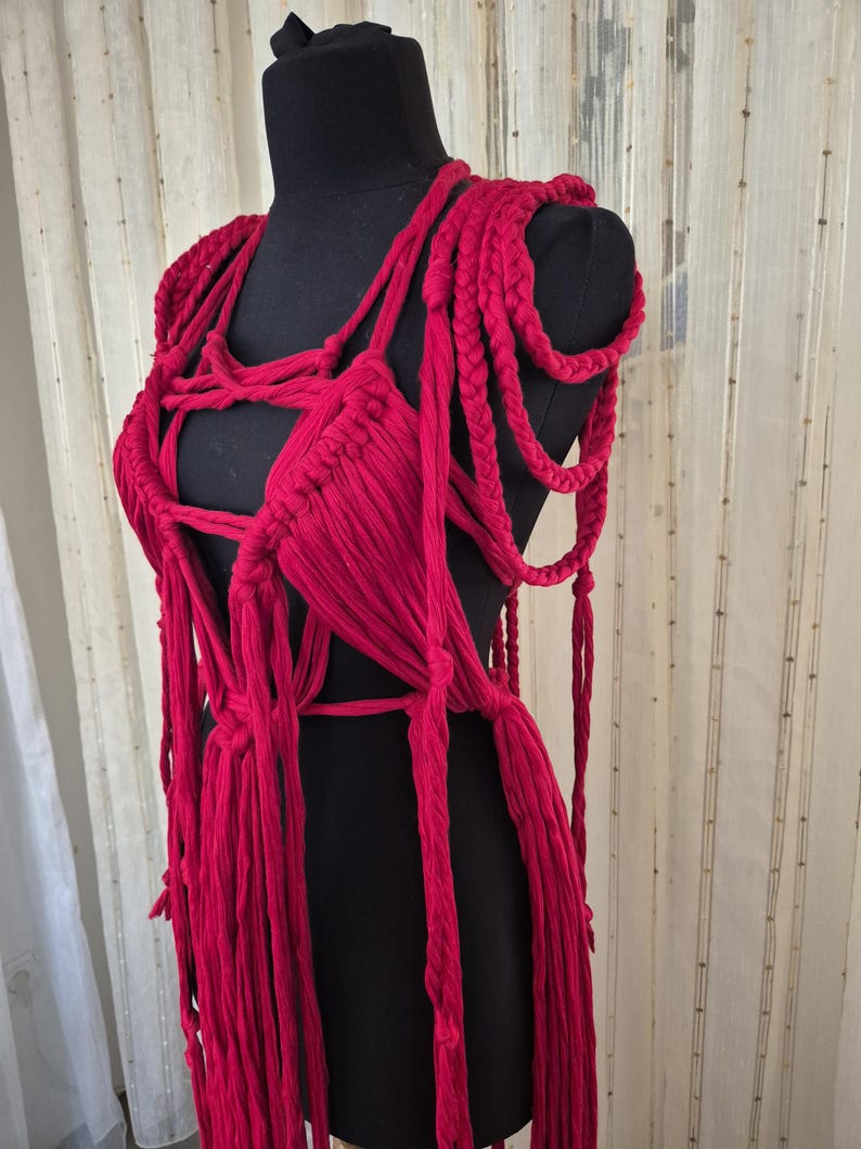 May include: A vibrant red macrame garment featuring intricate hand-knotted cord patterns, thick braided shoulder straps, and long dangling fringe elements. The piece is constructed from soft fabric strips, creating a complex, open-weave structure.