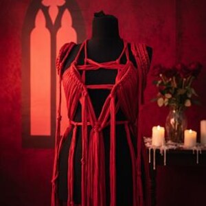 May include: A red macrame garment featuring a complex arrangement of knotted fabric cords that form a structured bodice with a pentagram-like design at the chest, extending into long, flowing vertical fringe strands that drape down from the waist.