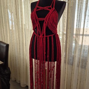 May include: A deep red macrame garment featuring a halter-style neckline with intricate knotted cord detailing across the chest and a long, flowing fringe that extends downward from the waist, displayed on a black mannequin torso.