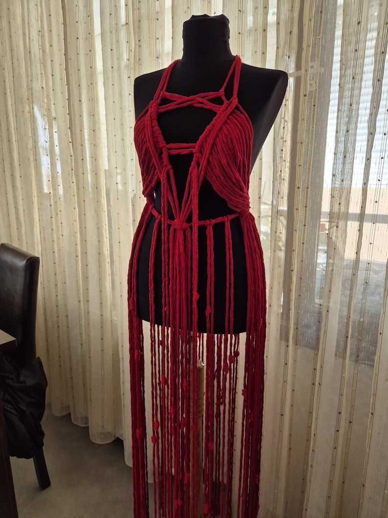 May include: A deep red macrame garment featuring a halter-style neckline with intricate knotted cord detailing across the chest and a long, flowing fringe that extends downward from the waist, displayed on a black mannequin torso.