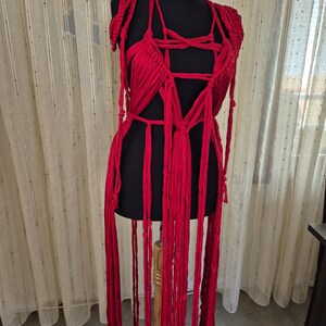 May include: A bright red macrame garment featuring a complex arrangement of knotted fabric strips, a deep V-neckline, and long, dangling fringe elements that extend downward from the waist, displayed on a black mannequin torso.
