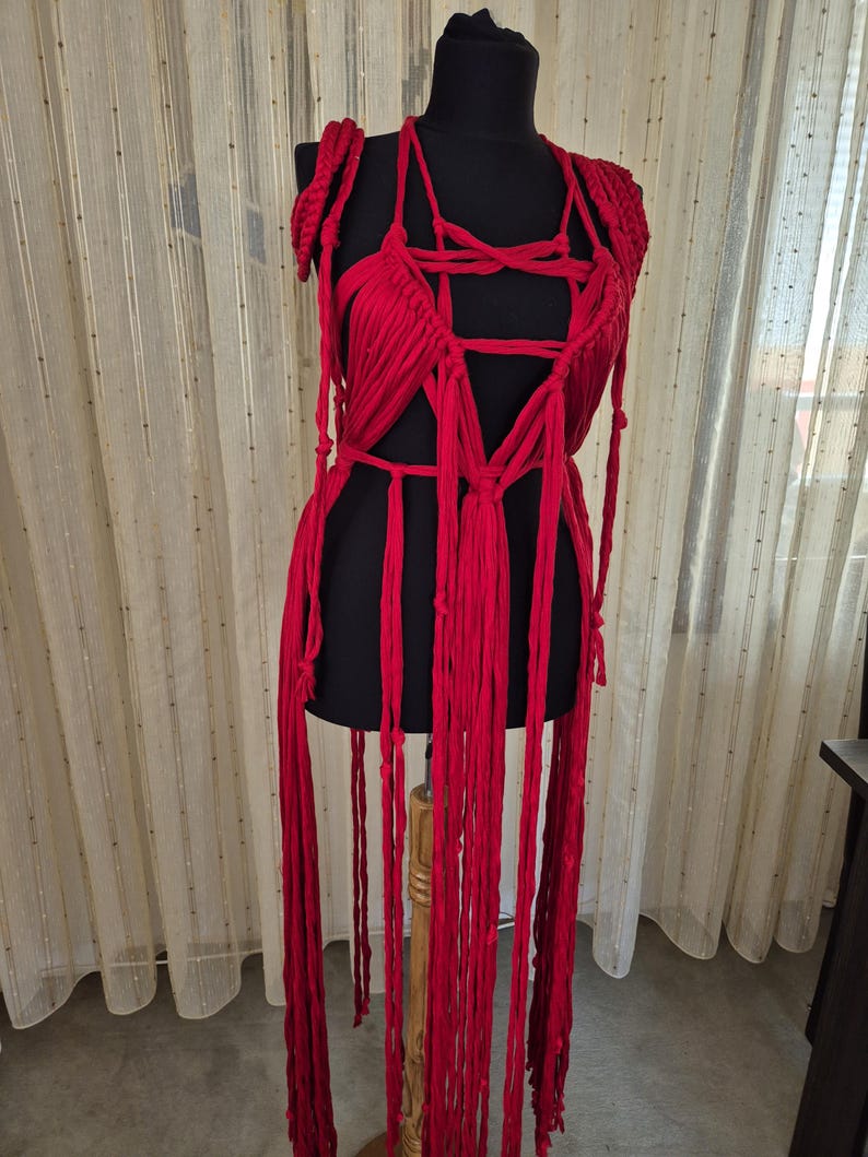 May include: A bright red macrame garment featuring a complex arrangement of knotted fabric strips, a deep V-neckline, and long, dangling fringe elements that extend downward from the waist, displayed on a black mannequin torso.
