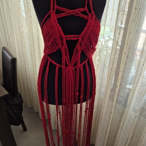 May include: A bright red macrame halter top garment featuring intricate knotted cord patterns, a deep neckline, and long, dangling fringe strands that extend downward, displayed on a black mannequin torso against a sheer curtain background.