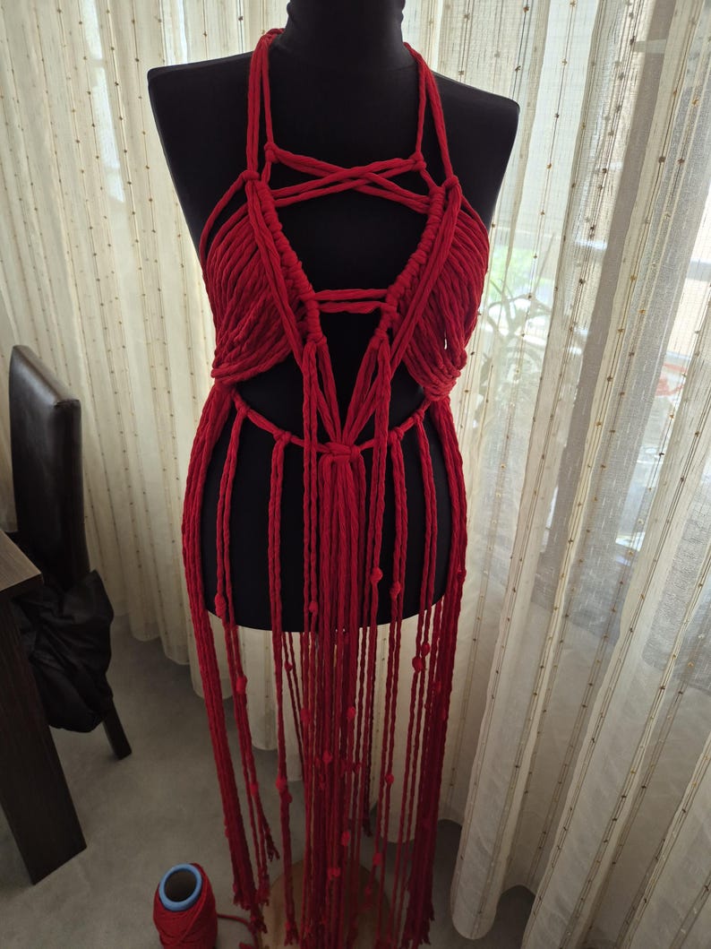May include: A bright red macrame halter top garment featuring intricate knotted cord patterns, a deep neckline, and long, dangling fringe strands that extend downward, displayed on a black mannequin torso against a sheer curtain background.