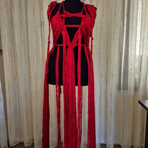 May include: A bright red garment constructed from multiple long fabric strips and knotted cords. The piece features a complex arrangement of vertical strands and woven sections that drape downward, creating a layered, fringe-like appearance.