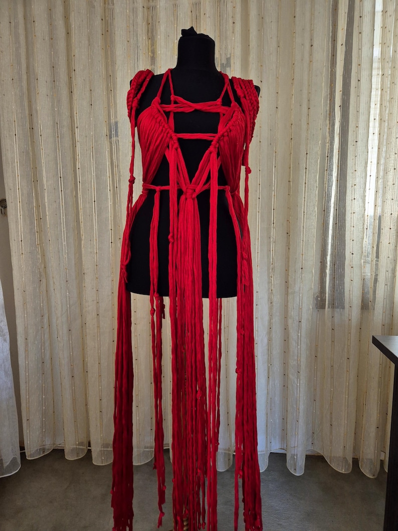 May include: A bright red garment constructed from multiple long fabric strips and knotted cords. The piece features a complex arrangement of vertical strands and woven sections that drape downward, creating a layered, fringe-like appearance.