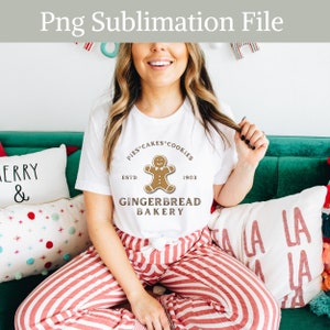 May include: A white t-shirt with a gingerbread man graphic and the text "PIES CAKES COOKIES ESTD 1903 GINGERBREAD BAKERY". The person wearing the shirt is sitting on a couch with red and white striped pants.