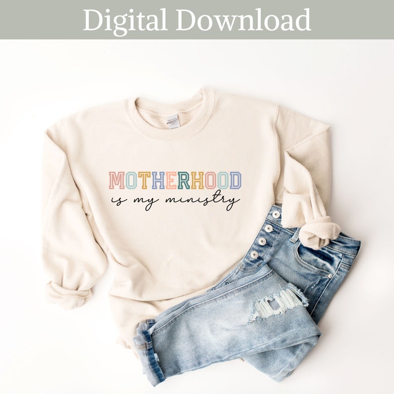 May include: A beige sweatshirt with the text "MOTHERHOOD is my ministry" in rainbow colors. The sweatshirt is folded and laid on a white surface with a pair of blue jeans.