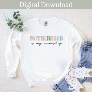 May include: White sweatshirt with the text "MOTHERHOOD is my ministry" in colorful outlined letters. The letters are in shades of pink, yellow, blue, and green.