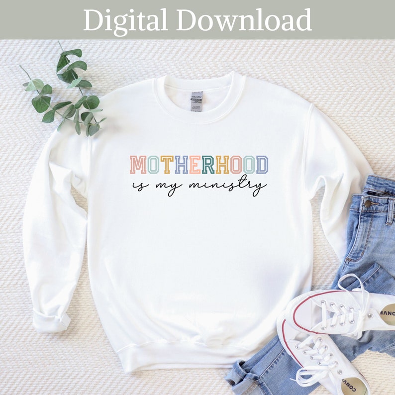 May include: White sweatshirt with the text "MOTHERHOOD is my ministry" in colorful outlined letters. The letters are in shades of pink, yellow, blue, and green.
