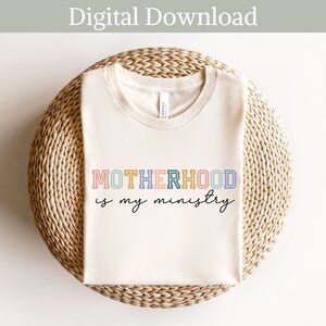 May include: A white t-shirt with the text "MOTHERHOOD is my ministry" in colorful letters. The letters are outlined in black and have a retro, vintage look.