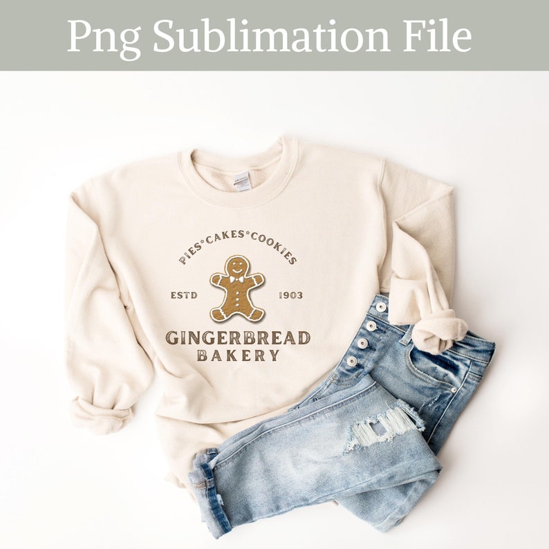 May include: A beige sweatshirt with a gingerbread man graphic and the text "Pies Cakes Cookies Est'd 1903 Gingerbread Bakery". The sweatshirt is folded and placed on top of a pair of blue jeans.