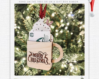 Könnte beinhalten: Ein hölzerner Weihnachtsbaumschmuck in Form einer Tasse mit einem Geschenkkartenhalter. Die Tasse hat einen weißen Schlagsahne-Deckel mit roten und grünen Streuseln und einem roten und weißen Zuckerstangen. Die Tasse sagt "Frohe Weihnachten" und hat auf jeder Seite ein Rentier. Der Schmuck hängt an einem Weihnachtsbaum.