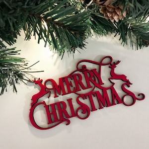 Puede incluir: Decoración navideña de madera roja con las palabras "MERRY CHRISTMAS" en una fuente decorativa. Las letras están conectadas y presentan siluetas de renos. Ramas de pino verdes y una piña en el fondo.