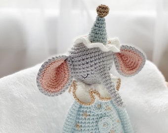 May include: A crocheted stuffed elephant wearing a blue and white hat with a gold star on top. The elephant is wearing a blue and white outfit with gold stars and a gold trim. The elephant is sitting on a white blanket.