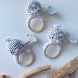 May include: Three crocheted whale teethers with wooden rings. The whales are gray and white with black eyes. The teethers are on a white background with a piece of driftwood.