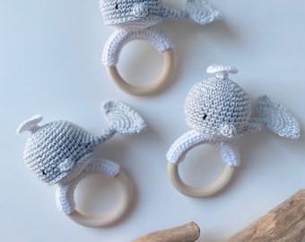 May include: Three crocheted whale teethers with wooden rings. The whales are gray and white with black eyes. The teethers are on a white background with a piece of driftwood.