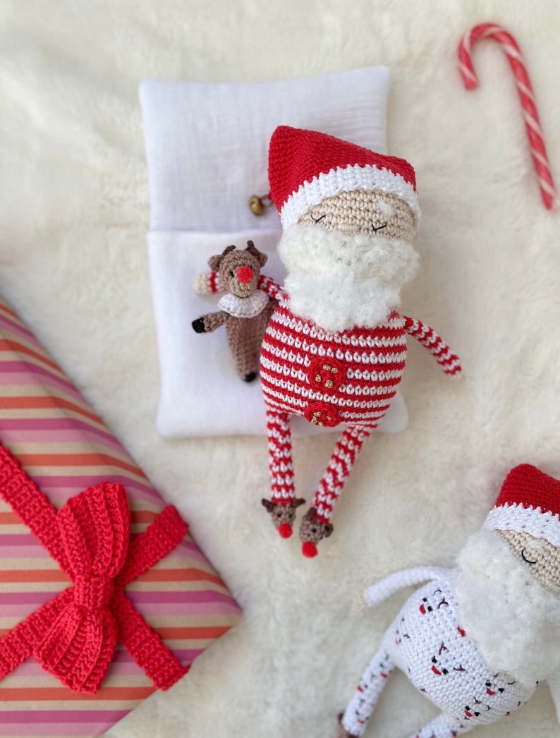 May include: Two crocheted Santa Claus dolls in red and white pajamas. One Santa is lying down on a white pillow with a crocheted reindeer doll. The other Santa is standing up with a white and red striped pajama top and a white pajama bottom with a red and white polka dot pattern.