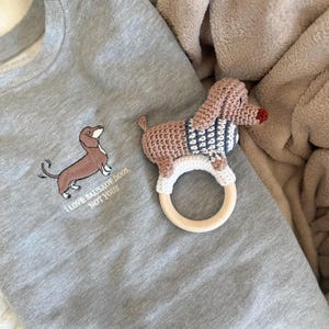 Op de afbeelding: Een grijze sweatshirt met een geborduurde teckel en de tekst "I LOVE SAUSAGE DOGS, NOT YOU!!!". Daarnaast een gehaakte teckel rammelaar met een houten ring. De rammelaar heeft een roze lichaam, een blauw-witte trui en een rode neus.