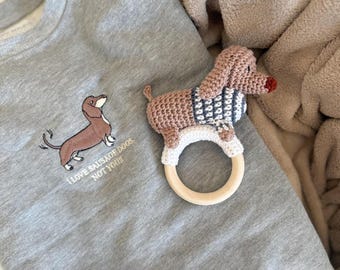 May include: A gray sweatshirt with an embroidered dachshund and the text "I LOVE SAUSAGE DOGS, NOT YOU!!!" A crocheted dachshund rattle with a wooden ring is next to the sweatshirt. The rattle has a pink body, blue and white sweater, and a red nose.