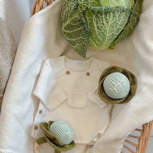 Op de afbeelding: Een witte baby romper met een zakje en twee gehaakte kool rammelaars. De rammelaars zijn groen en hebben een houten ring. De romper heeft het woord "soulflow" op het label.