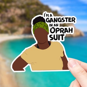 Pode incluir: Um adesivo com uma ilustração de desenho animado de uma pessoa de pele escura vestindo uma camisa amarela e uma faixa de cabeça verde. O texto no adesivo diz "I'm a gangster in an Oprah suit."