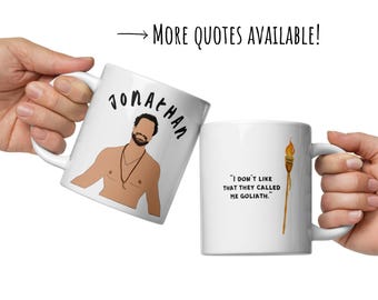 May include: Two white ceramic mugs. One mug features a cartoon illustration of a man with the name "Jonathan" above. The other mug has the quote, "I don't like that they called me Goliath." and a torch illustration.