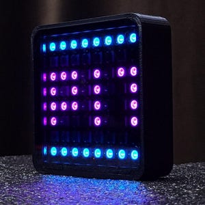 以下が含まれることがあります： 青とピンクのLEDライトで「Hi」という文字を表示する黒い正方形のデジタルディスプレイ。デバイスは暗い仕上げで、テクスチャ加工された表面にあります。ライトはグリッドパターンに配置されています。