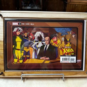 Op de afbeelding: Een ingelijste comicboekomslag met de X-Men-personages. De omslag is in een houten lijst met een bruin en wit houtnerfpatroon. De comicboekomslag is getiteld "X-Men '97" en toont de personages Storm, Professor X, Wolverine, Cyclops, Jean Grey en Rogue. De comicboekomslag is in een bruin en gouden kleurenschema.