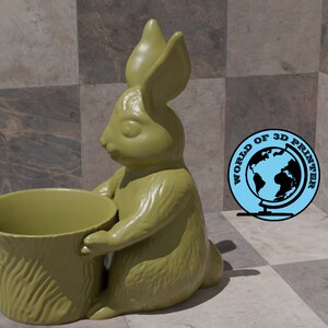 May include: A green ceramic rabbit planter holding a green ceramic pot with a wood-like texture. The rabbit is sitting on a checkered surface.