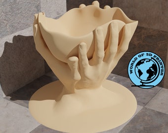 Op de afbeelding: Een beige keramische sculptuur van twee handen die een kom vasthouden. De sculptuur staat op een beige basis en staat op een betegelde ondergrond.
