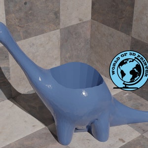 Puede incluir: Un macetero de dinosaurio de color azul claro con una abertura ancha en la parte superior. El macetero está hecho de plástico y tiene un acabado liso y brillante.