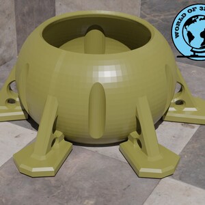 Puede incluir: Un objeto en forma de cúpula impreso en 3D, de color verde oliva, con un centro hueco y cuatro patas. El objeto tiene una superficie lisa y redondeada y una base ligeramente texturizada.