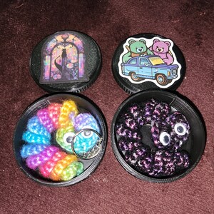 May include: Four round black containers with lids. Each container has a different design on the lid. One lid has a stained glass window design, another has a cartoon of two bears in a car, and the other two containers have colorful crocheted items inside.